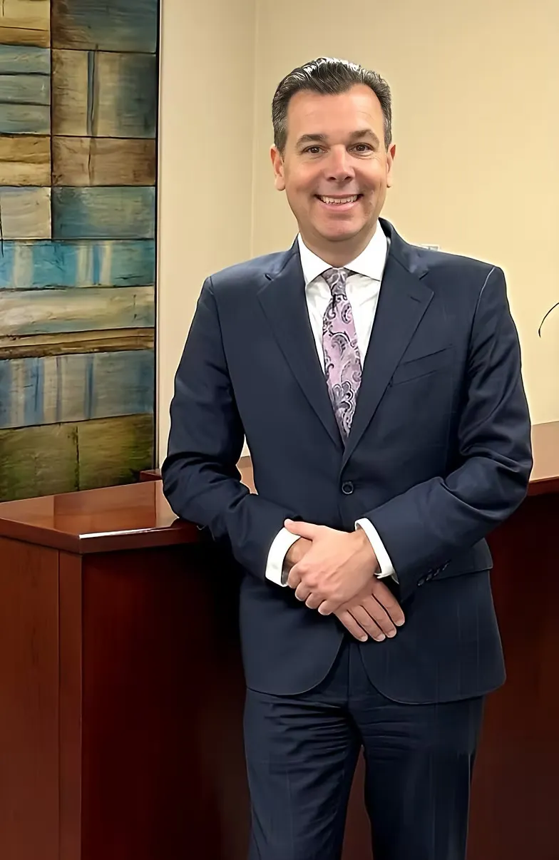 Professional man in navy blue suit and patterned tie standing indoors by wooden desk.