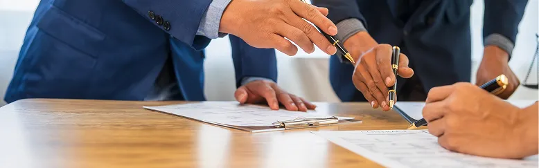 Three individuals in business attire reviewing and signing documents on a wooden table.