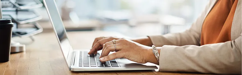 Person wearing beige jacket typing on silver laptop at wooden desk with black coffee cup.