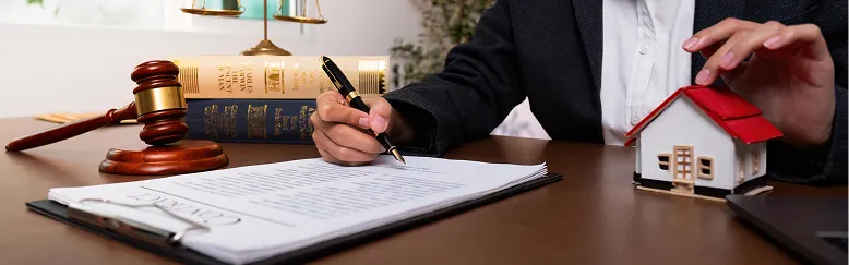 Person signing legal documents at desk with gavel, law books, and miniature house model.