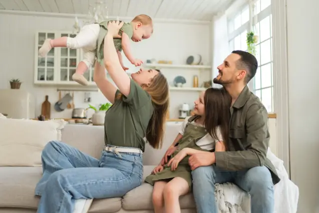 A family of four in a bright living room, with a mother lifting a baby while the father and daughter watch smiling.